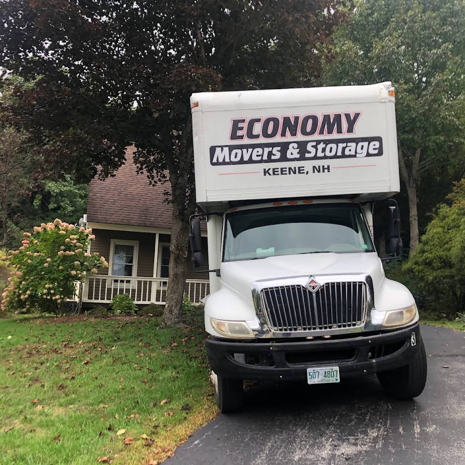 Economy Movers truck sitting in driveway of a small brown house with trees and green grass in the background